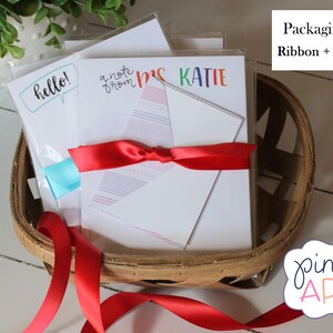 May include: A set of three blank note cards with different designs, including "hello!" in a speech bubble, "a note from" with a rainbow design, and "Mrs. Katie" in rainbow letters. The cards are wrapped in a basket with a red ribbon and a gift message.