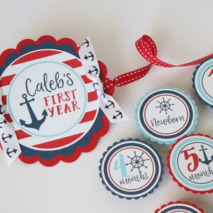 Może przedstawiać: Baner z kamieniami milowymi dla dziecka w stylu marynistycznym w kolorze czerwonym, białym i niebieskim z napisem "Caleb's First Year" i kotwicą. Baner zawiera 12 osobnych k&oacute;łek z numerami i napisem "month" lub "months".