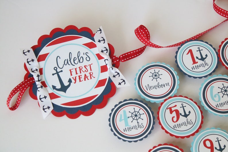 Może przedstawiać: Baner z kamieniami milowymi dla dziecka w stylu marynistycznym w kolorze czerwonym, białym i niebieskim z napisem "Caleb's First Year" i kotwicą. Baner zawiera 12 osobnych k&oacute;łek z numerami i napisem "month" lub "months".