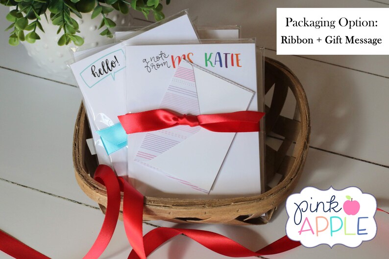 May include: A set of three blank note cards with different designs, including "hello!" in a speech bubble, "a note from" with a rainbow design, and "Mrs. Katie" in rainbow letters. The cards are wrapped in a basket with a red ribbon and a gift message.