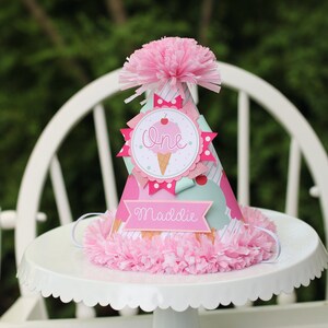 May include: A pink and white ice cream cone party hat with the word "one" and the name "Maddie" on it. The hat is decorated with pink and white polka dots, a pink bow, and a pink cherry.