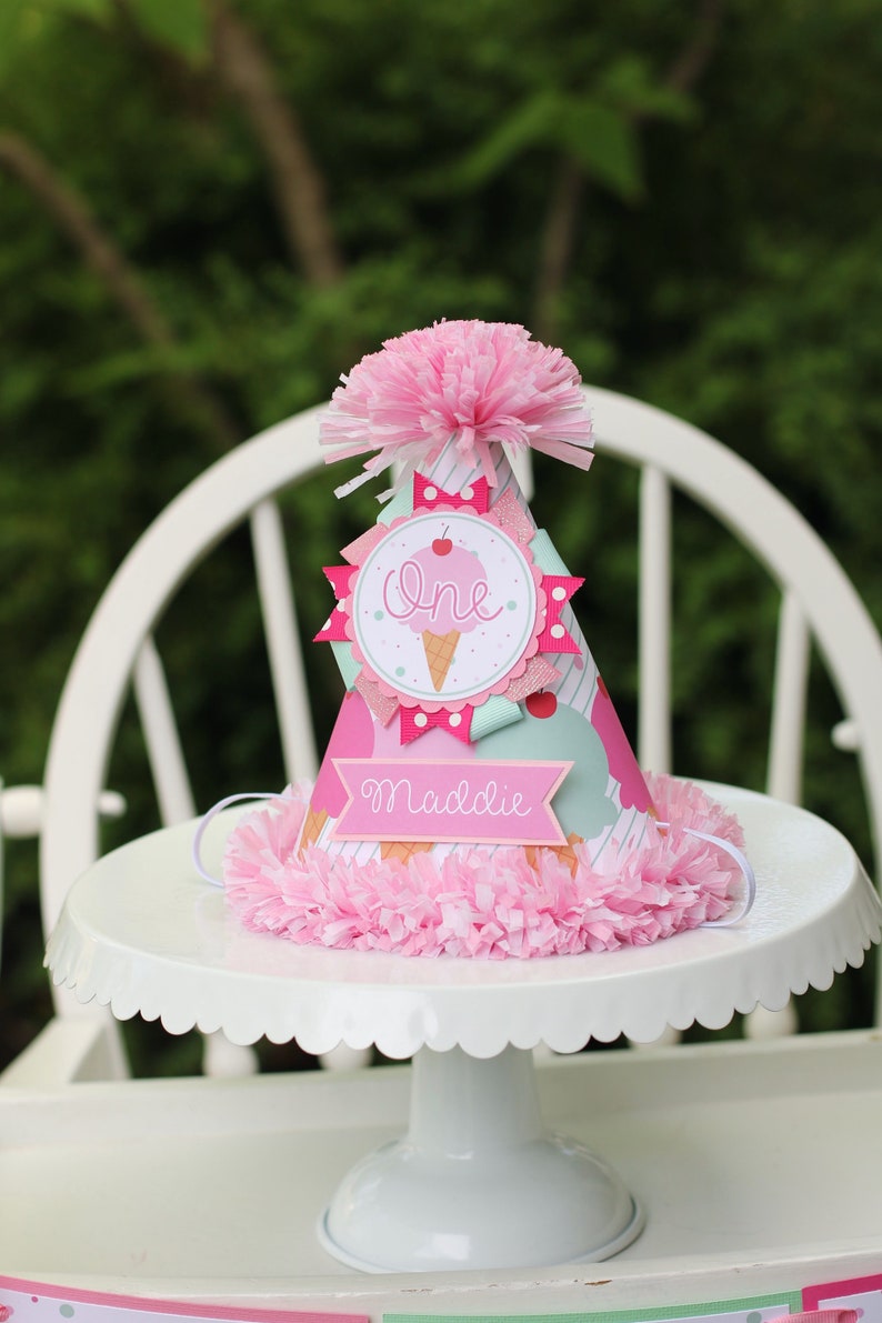 May include: A pink and white ice cream cone party hat with the word "one" and the name "Maddie" on it. The hat is decorated with pink and white polka dots, a pink bow, and a pink cherry.