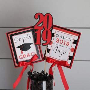 May include: Graduation centerpiece with red, black, and white decorations. The centerpiece includes a graduation cap, the words "Congrats Grad", and the numbers "2019". The other sign says "Class of 2019 Angie".
