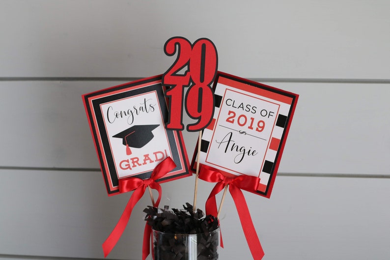 May include: Graduation centerpiece with red, black, and white decorations. The centerpiece includes a graduation cap, the words "Congrats Grad", and the numbers "2019". The other sign says "Class of 2019 Angie".