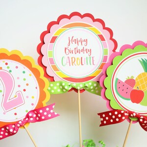 May include: Three colorful round cake toppers with scalloped edges. The toppers feature a pink number "2", a striped design with the text "Happy Birthday Caroline", and a fruit design with a watermelon, pineapple, apple, pear, and orange. Each topper has a red and white polka dot ribbon.