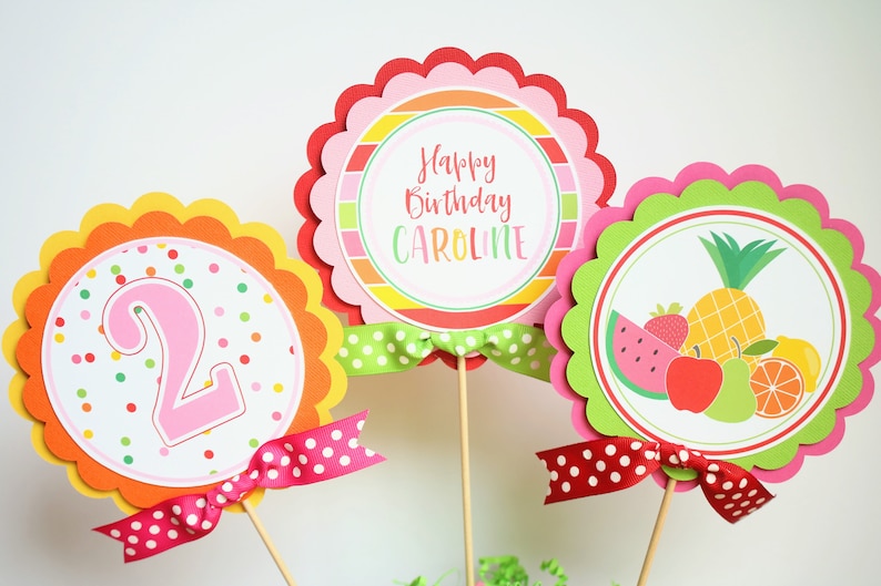 May include: Three colorful round cake toppers with scalloped edges. The toppers feature a pink number "2", a striped design with the text "Happy Birthday Caroline", and a fruit design with a watermelon, pineapple, apple, pear, and orange. Each topper has a red and white polka dot ribbon.