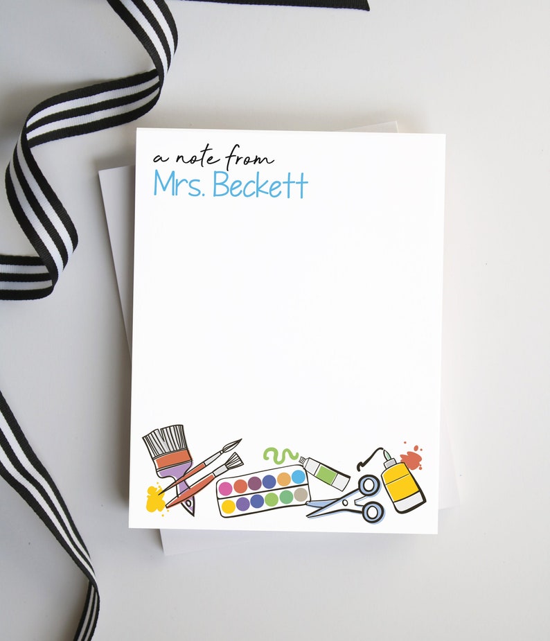 May include: A white notecard with the text "a note from Mrs. Beckett" in black and blue lettering. The bottom of the card features a colorful illustration of art supplies, including paintbrushes, a paint palette, scissors, and glue.