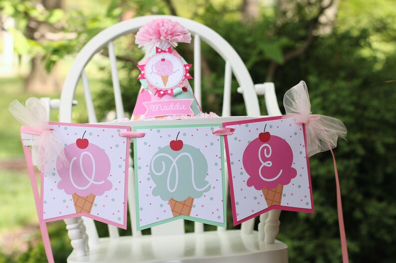 May include: A pink and green ice cream cone themed banner with the word "ONE" spelled out in pink and green letters. The banner is decorated with pink and white polka dots and cherries. The banner is hanging on a white high chair with a pink and white ice cream cone themed party hat on top.