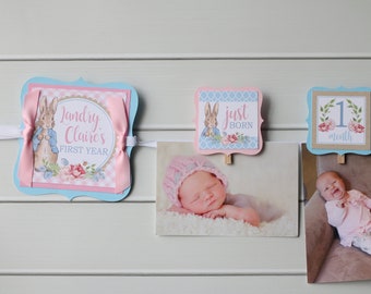 May include: A baby photo banner with a Peter Rabbit theme. The banner includes a photo of a newborn baby and a photo of a baby smiling. The banner has a blue and pink color scheme with floral accents and the text "Jandry Claire's FIRST YEAR".