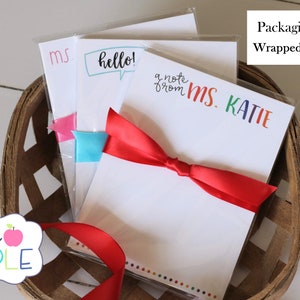May include: Three packs of white notepad paper with colorful text, wrapped with a red ribbon. The text on the notepads reads "ms", "hello!" and "a note from MS. KATIE".
