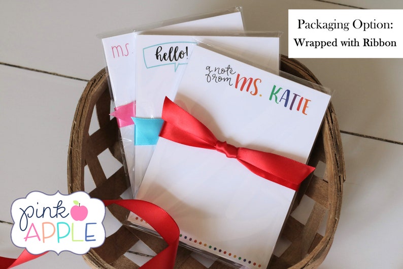 May include: Three packs of white notepad paper with colorful text, wrapped with a red ribbon. The text on the notepads reads "ms", "hello!" and "a note from MS. KATIE".