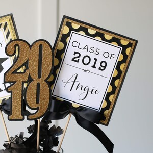 Puede incluir: Centro de mesa de graduaci&oacute;n con decoraciones negras y doradas. El centro de mesa incluye un letrero que dice "Clase de 2019 Angie", un letrero que dice "Felicidades Graduado", y un letrero "2019" de purpurina dorada.