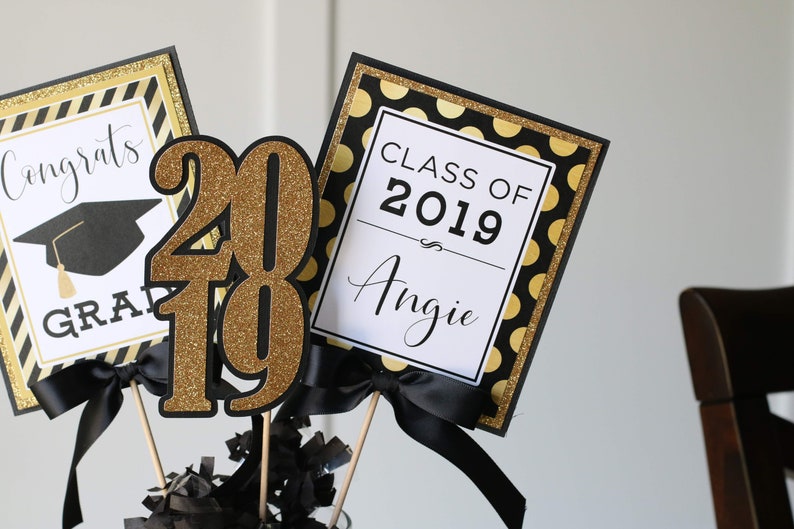 Puede incluir: Centro de mesa de graduaci&oacute;n con decoraciones negras y doradas. El centro de mesa incluye un letrero que dice "Clase de 2019 Angie", un letrero que dice "Felicidades Graduado", y un letrero "2019" de purpurina dorada.