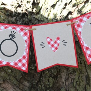 May include: Red and white gingham banner with the words "To" and "Be" and a ring with a diamond in the centre. The banner is strung with twine.
