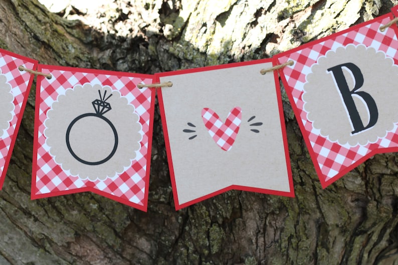 May include: Red and white gingham banner with the words "To" and "Be" and a ring with a diamond in the centre. The banner is strung with twine.