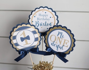 May include: Three blue and gold glitter paper circles with scalloped edges. The top circle says "Happy Birthday Brixton". The left circle says "1" with a gold bow tie. The right circle says "Mr. ONEderful".
