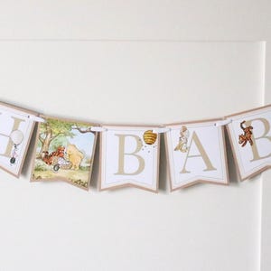 May include: A Winnie the Pooh themed baby banner with the letters "Oh Baby" in gold and white. Each letter is on a separate pennant with a Winnie the Pooh character illustration. The banner is strung with white ribbon.