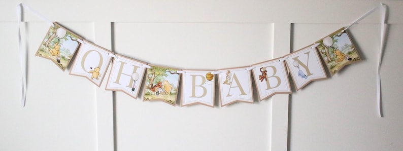 May include: A Winnie the Pooh themed baby banner with the letters "Oh Baby" in gold and white. Each letter is on a separate pennant with a Winnie the Pooh character illustration. The banner is strung with white ribbon.