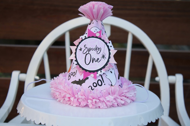 May include: A pink and white Halloween-themed party hat with the words "Spooky One" and "Boo!" printed on it. The hat features a spider web design, a spider, and ghost illustrations. It is decorated with pink tissue paper.