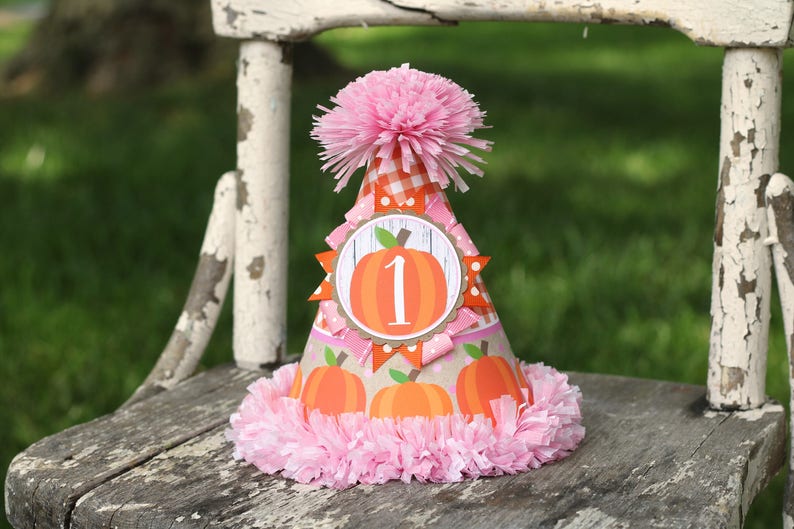 May include: A pink and orange pumpkin-themed party hat with a pink pom-pom on top. The hat has a white circle with a number "1" and a pumpkin inside. The hat is sitting on a wooden chair.