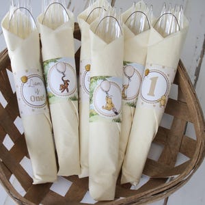 May include: A woven basket holds several cream-colored napkins, each wrapped around clear plastic cutlery. The napkins are decorated with Winnie the Pooh themed stickers, some with the number 1, and others with the text "Liam is One!"