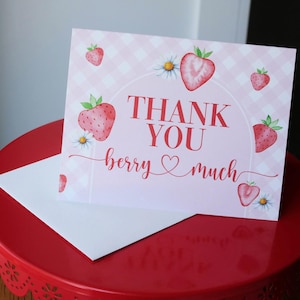 May include: A pink and white card with the words "THANK YOU berry much" in red script, decorated with strawberry and daisy illustrations. A white envelope rests below the card on a red cake stand.