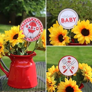 Op de afbeelding: Een rode keramische kan gevuld met zonnebloemen, met decoratieve borden met de tekst "BABY Q" en "Welcome BABY". De borden zijn rond met een rood-wit geblokt patroon en baby-thema graphics. De zonnebloemen zijn geel.