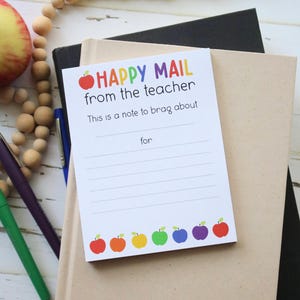 May include: A white card with the text "Happy Mail from the teacher. This is a note to brag about for" and a red apple icon. The bottom of the card has a row of rainbow colored apples.