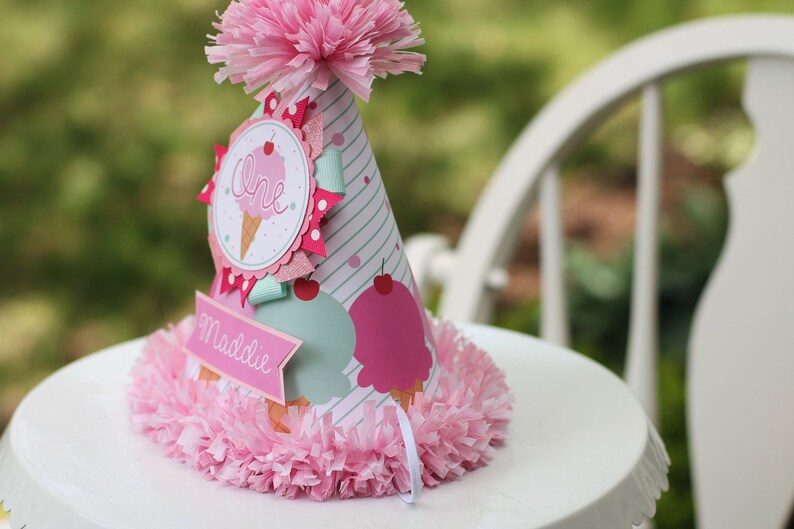 May include: A pink and blue ice cream cone party hat with the word "One" and the name "Maddie" on it. The hat is decorated with pink and white tissue paper.