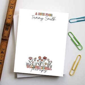 May include: A white notepad with the text "A Note From Tammy Smith" at the top and "Occupational Therapy" with floral illustrations at the bottom. A wooden ruler and colorful paper clips are in the background.