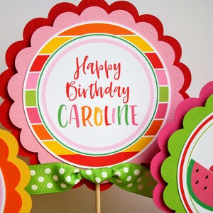 May include: A set of three colorful round cake toppers with scalloped edges. The center topper features a pink and white striped background with the text "Happy Birthday Caroline". The other two toppers feature a fruit theme with a watermelon, pineapple, strawberry, and apple.