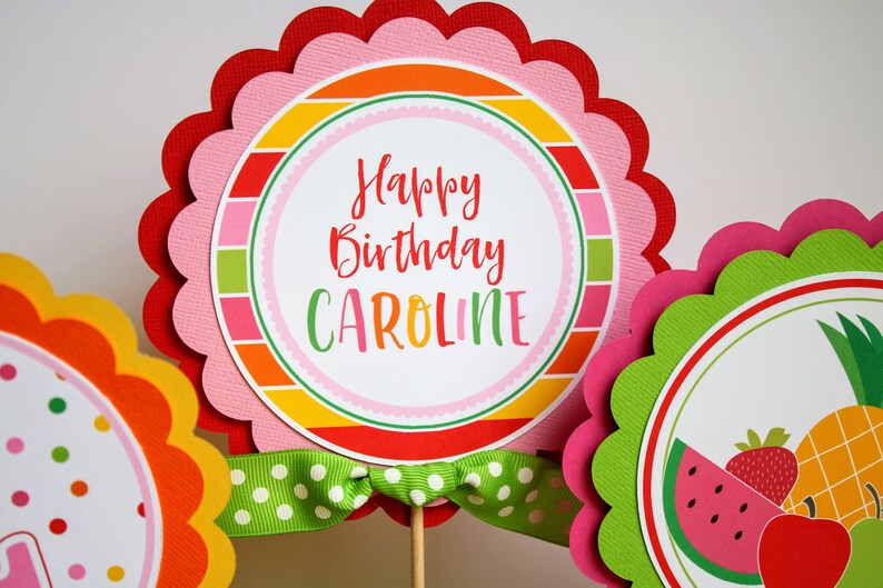 May include: A set of three colorful round cake toppers with scalloped edges. The center topper features a pink and white striped background with the text "Happy Birthday Caroline". The other two toppers feature a fruit theme with a watermelon, pineapple, strawberry, and apple.