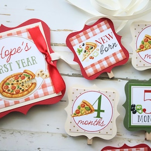 May include: A collection of decorative signs with a pizza theme. One sign reads "Hope's FIRST YEAR" with a pizza illustration. Other signs mark "New Born", "1 month", "4 months", and "5 months". The signs have red, green, and beige accents.
