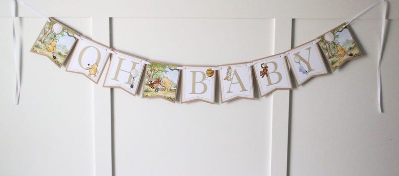 May include: A Winnie the Pooh themed baby shower banner with the letters "OH BABY" in gold lettering on a white background. Each letter is decorated with a Winnie the Pooh character.