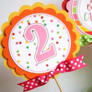 May include: A pink number two with colorful polka dots on a white background. The number is on a scalloped orange and yellow circle with a pink and white polka dot ribbon.
