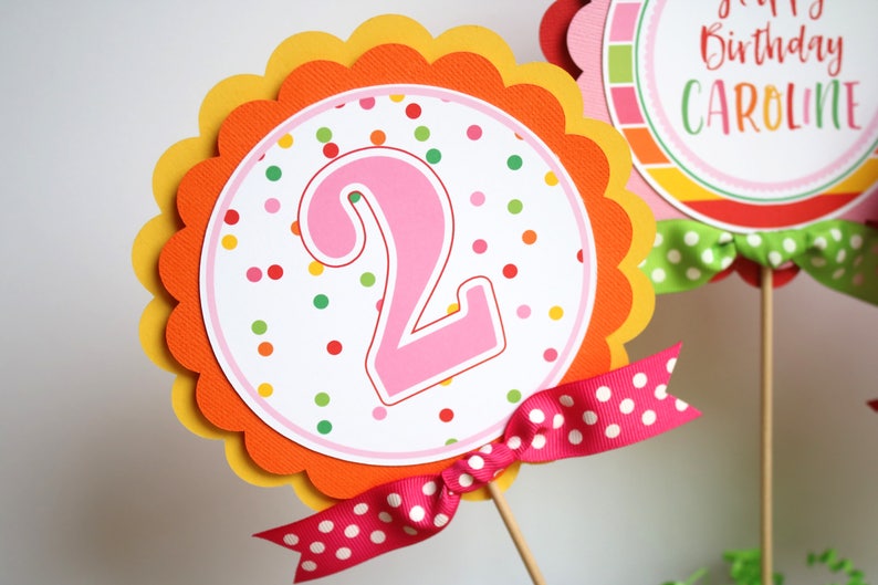May include: A pink number two with colorful polka dots on a white background. The number is on a scalloped orange and yellow circle with a pink and white polka dot ribbon.