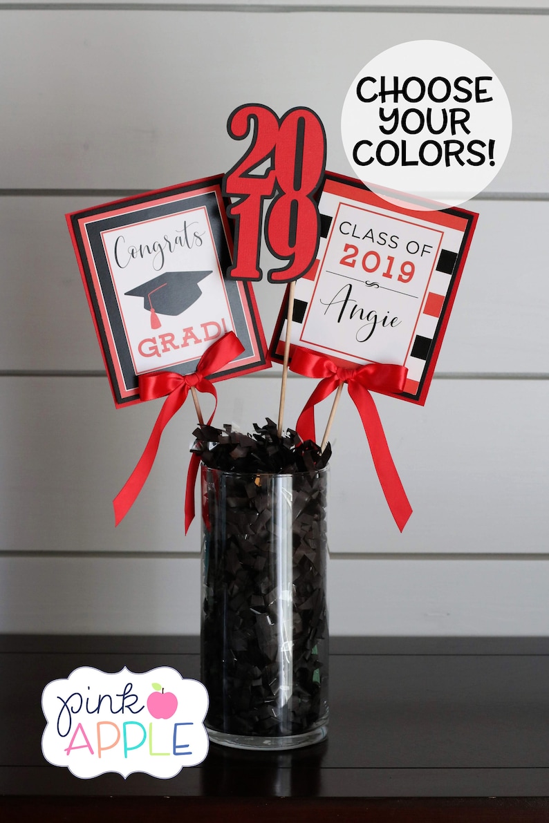 May include: Graduation centerpiece with three red and black signs. The signs say "Congrats Grad", "Class of 2019 Angie", and "2019". The signs are in a clear vase with black shredded paper.