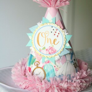 May include: A pink and white party hat with a blue and white ribbon and a gold circle with the word "One" in gold lettering. The hat is decorated with a pink and white checkered pattern and a pink pom-pom on top.