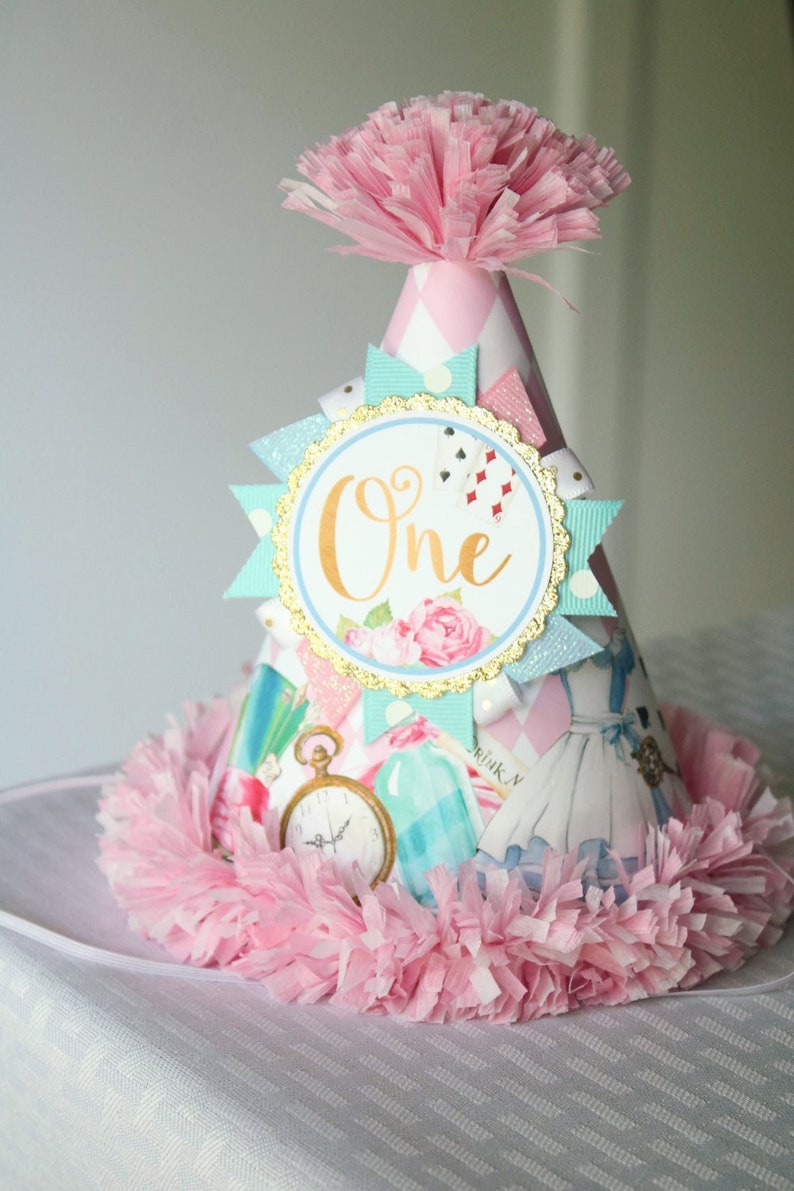 May include: A pink and white party hat with a blue and white ribbon and a gold circle with the word "One" in gold lettering. The hat is decorated with a pink and white checkered pattern and a pink pom-pom on top.