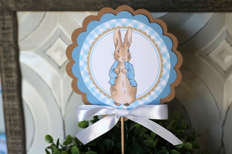 K&ouml;nnte beinhalten: Dekorative Tortenaufsatz mit Peter Rabbit in blauem Jackett, vor blau-wei&szlig;em Vichy-Muster. Der Aufsatz hat einen gewellten Rand und ist mit einer wei&szlig;en Schleife an einem Holzstab befestigt. Ideal f&uuml;r eine Babyparty oder Ostern.