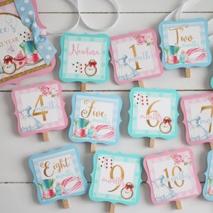 May include: Alice's First Year monthly milestone markers.  Each card features a different Alice in Wonderland theme with a number and the word 'months' in gold lettering.  The cards are pink and blue with a white background.