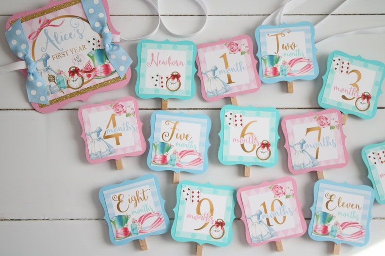 May include: Alice's First Year monthly milestone markers.  Each card features a different Alice in Wonderland theme with a number and the word 'months' in gold lettering.  The cards are pink and blue with a white background.