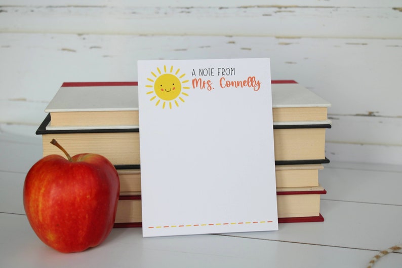 Puede incluir: Un bloc de notas blanco con un sol amarillo sonriente y el texto "A note from Mrs. Connelly" en naranja. El bloc de notas est&aacute; sobre una pila de libros con una manzana roja delante.
