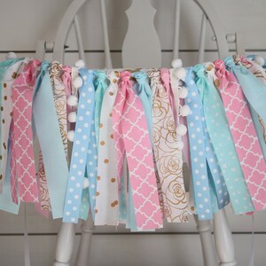 May include: A pink, blue, and white fabric banner with gold accents and white pom-poms. The banner is hanging on a white chair.