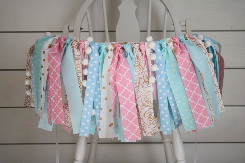 May include: A pink, blue, and white fabric banner with gold accents and white pom-poms. The banner is hanging on a white chair.