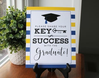 May include: A white sign with blue and yellow stripes. The sign says "Please share your key to success with the graduate!" with a black graduation cap and a key graphic.
