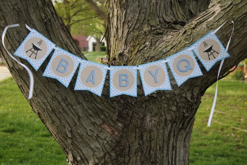 May include: A blue and brown banner with the words "BABY Q" and a grill icon on each end. The banner is hanging from a tree branch.