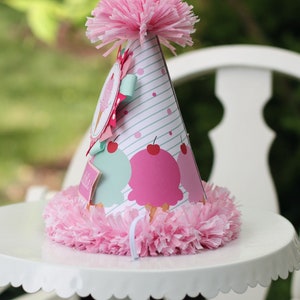 May include: A pink and white party hat with an ice cream cone design. The hat has a pink pom-pom on top and a white ruffle around the bottom.