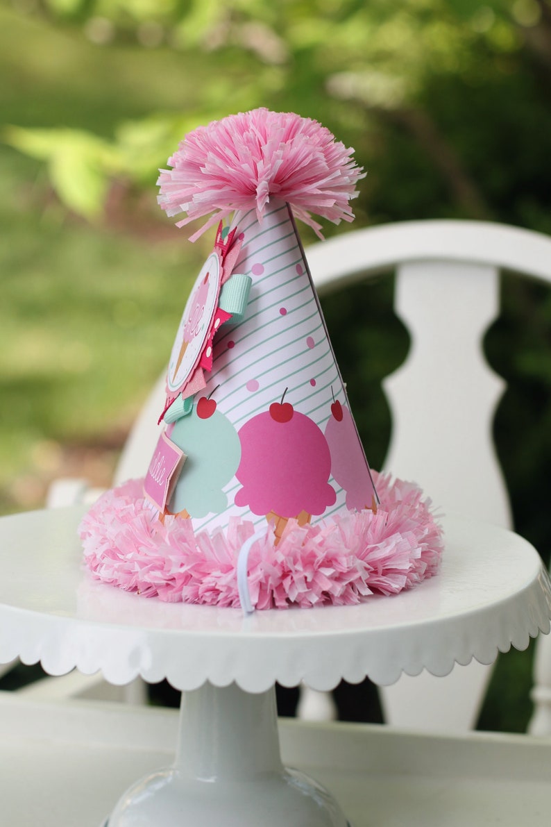 May include: A pink and white party hat with an ice cream cone design. The hat has a pink pom-pom on top and a white ruffle around the bottom.