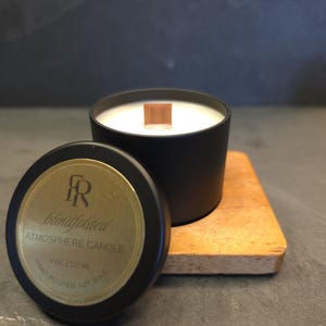 May include: A black metal candle tin with a wooden wick and white soy wax. The lid features a gold circular label with the text "FR blindfolded ATMOSPHERE CANDLE 4 OZ | 120 ML HAND POURED SOY WAX". The candle sits on a square wooden base.